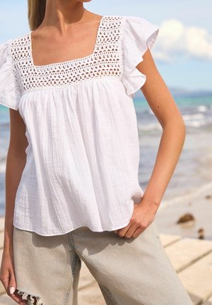 Blusa blanca de manga corta con escote cuadrado y detalles intrincados de crochet. Combinada con jeans claros y desgastados.