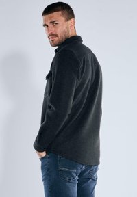 Mann mit kurzem Haar und Bart, der eine dunkelgraue Jacke und blaue Jeans trägt und über die Schulter gegen einen schlichten, hellen Hintergrund schaut.