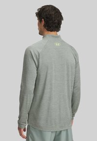Helle graue Langarm-Sportshirt mit hohem Kragen, strukturiertem Stoff und einem kleinen gelben Logo am oberen Rücken.