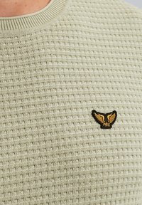 Jersey verde claro con patrón de punto texturizado y cuello acanalado. Presenta un logotipo de alas bordado en negro y dorado en el pecho.