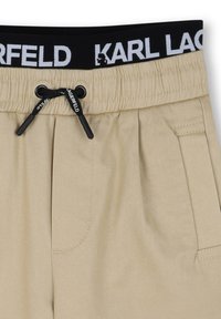 Beige broek met een zwarte elastische tailleband voorzien van witte tekst "KARL LAGERFELD", een trekkoord en zijzakken. Tedere textuur.