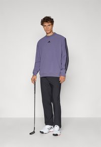 Pullover viola a maniche lunghe con strisce laterali nere, abbinato a pantaloni neri e scarpe da golf bianche, mentre tiene una mazza da golf.