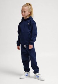 Marinblå hoodie med en framficka och en liten logotyp, tillsammans med matchande joggers, stiliserad med vita sneakers med blå accenter.