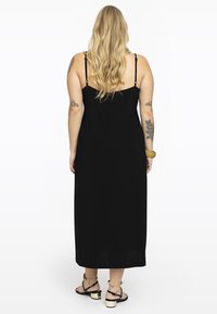 Schwarzes Maxikleid mit Spaghettiträgern, aus weichem Stoff und mit lässiger Passform. Es hat einen geraden Saum und einen subtilen Seitenschlitz. Wird mit hohen Sandalen kombiniert.