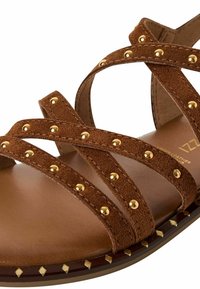 Bruine suède sandaal met gekruiste banden, versierd met gouden studs, een gewatteerde zool en een decoratieve rand van studs op de buitenzool.