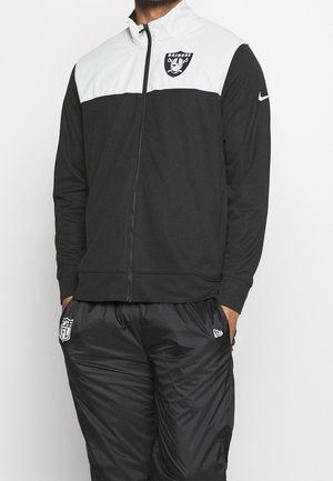 Personne portant une veste zippée noire et blanche Raiders et un pantalon de sport noir à la marque NFL, les mains dans les poches sur un fond uni.