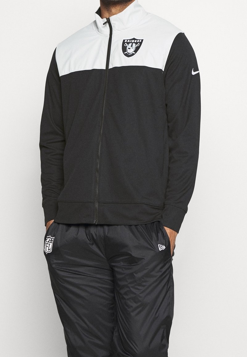 Personne portant une veste zippée noire et blanche Raiders et un pantalon de sport noir à la marque NFL, les mains dans les poches sur un fond uni.