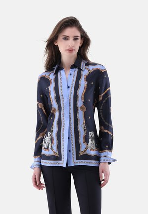 Donna che indossa una camicia botton-down blu navy e azzurra con stampe di catene e cani, abbinata a pantaloni neri, in piedi contro uno sfondo neutro.
