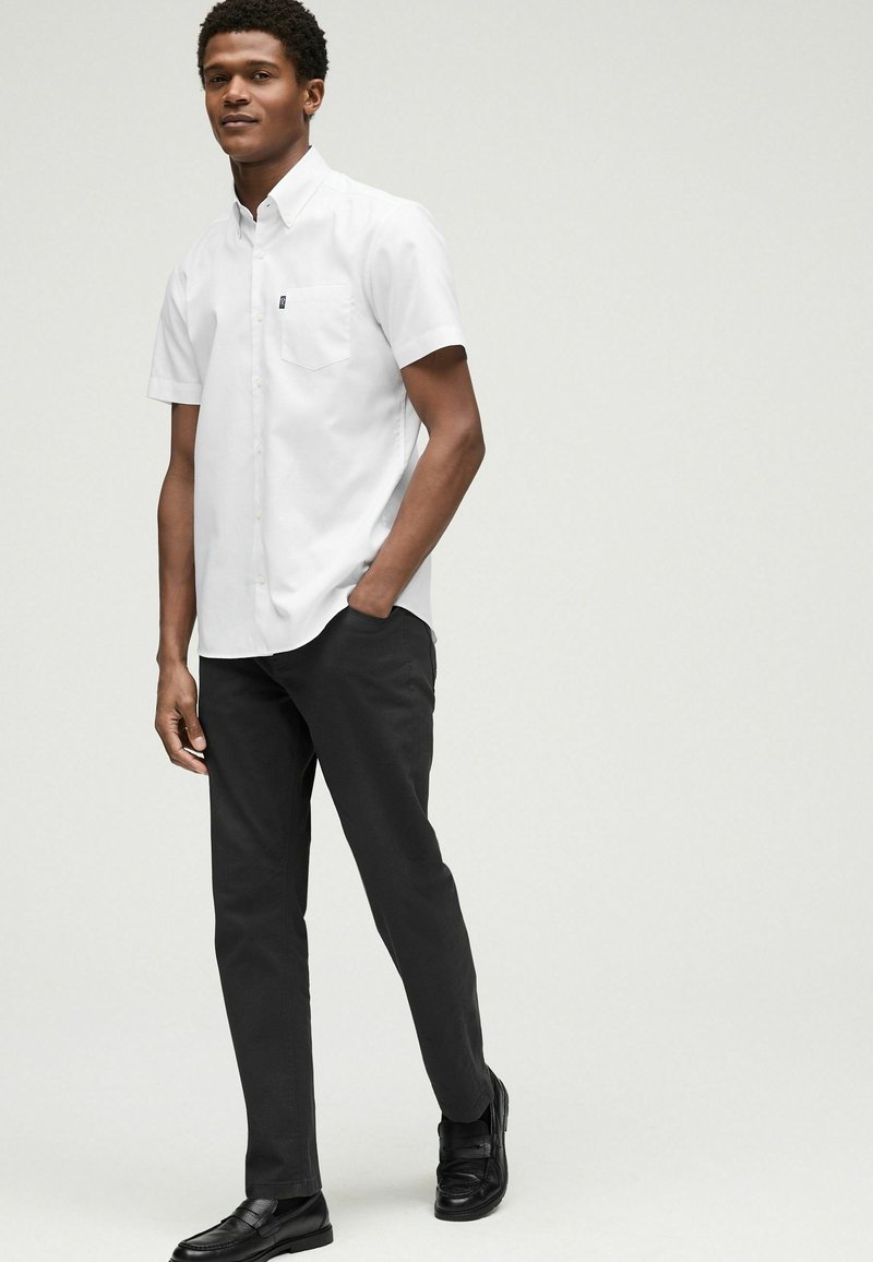 Weißes kurzärmliges Button-Down-Hemd mit einer Brusttasche, kombiniert mit dunklen Hosen und schwarzen Schuhen; glatte Textur und klassisches Design.