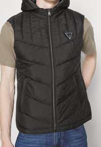 Schwarze Steppweste mit Kapuze, gestepptem Design, Vorderreißverschluss und Logopatch auf der Brust, getragen über einem beigen Hemd.