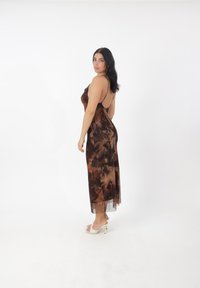 Robe glissante florale marron avec des bretelles fines et une superposition transparente, présentant une silhouette ajustée et un ourlet mi-long. Portée avec des talons clairs.