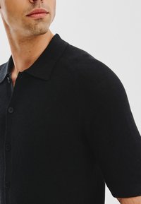 Mężczyzna w czarnej koszuli z krótkim rękawem i kołnierzykiem, lekko spoglądający w lewo na tle jasnego, jednolitego tła.