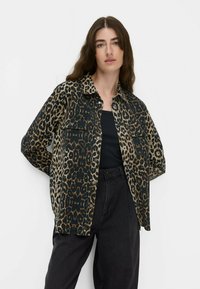 Eine Frau mit langen braunen Haaren, die ein knöpfenhemd mit Leopardenmuster, ein schwarzes Oberteil und lockere schwarze Hosen trägt, steht vor einem weißen Hintergrund.
