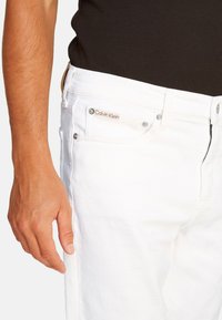 Jeans de mezclilla de algodón blanco con un corte ajustado, que presentan un logo de Calvin Klein en el bolsillo frontal, herrajes en tono plateado y cremallera en la bragueta.