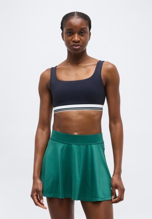 Athletic woman wearing a navy crop top with white stripe and a high-waisted green skater skirt, standing against a plain background.