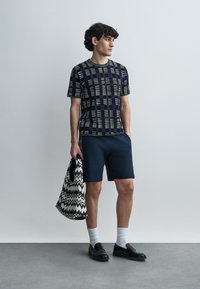 Κοντομάνικο πουκάμισο με σχέδιο σε ναυτικό μπλε, πράσινο και μπεζ, συνδυασμένο με σκούρα σορτς. Το μοντέλο κρατά μια μαύρη και λευκή τσάντα με ζιγκ ζαγκ και φορά μοκασίνια.