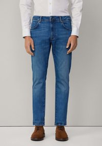 Vaqueros de mezclilla azul con corte recto, con cierre de botón, bolsillos laterales y costuras en contraste, combinados con zapatos de ante marrón.