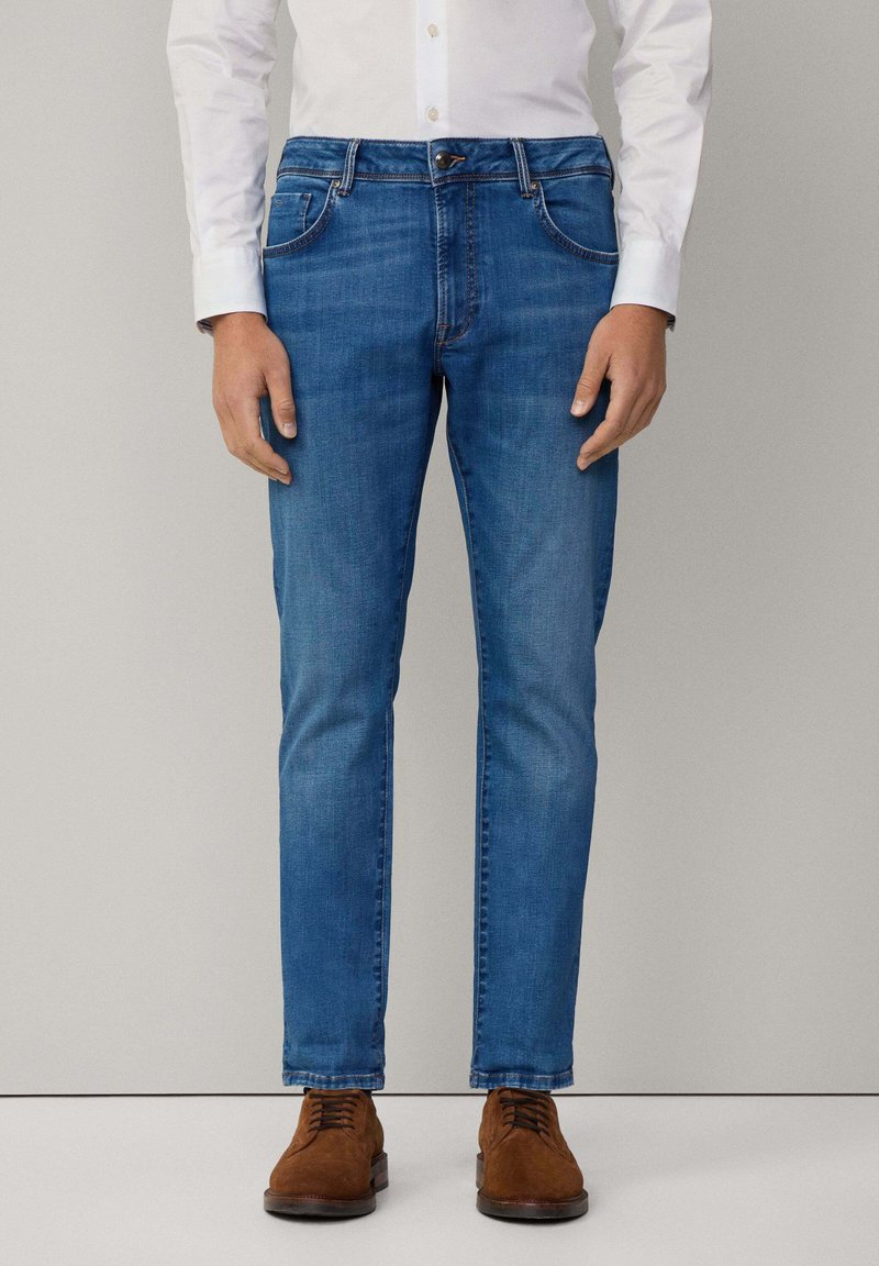 Vaqueros de mezclilla azul con corte recto, con cierre de botón, bolsillos laterales y costuras en contraste, combinados con zapatos de ante marrón.