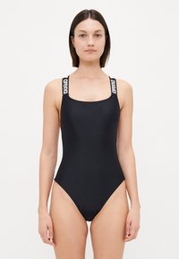Maillot de bain une pièce noir avec bretelles larges, détail de logo, décolleté carré, texture lisse et silhouette ajustée.