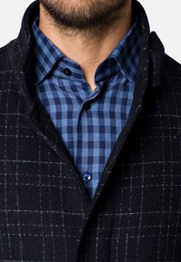 Czarny wzorzysty płaszcz o miękkiej fakturze na niebieskiej koszuli w kratę. Koszula ma ciemne guziki i klasyczny kołnierzyk.