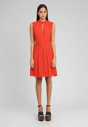 Jeune femme debout portant une robe rouge sans manches avec un décolleté en trou de serrure et des talons compensés noirs sur un fond gris uni.