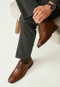 Chaussures habillées en cuir marron avec un bout pointu, un laçage à six oeillets, associées à un pantalon gris foncé et des chaussettes marron, sur un fond clair.