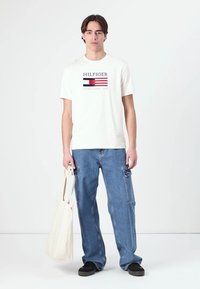 Bílý bavlněný tričko s logem "HILFIGER" a červenými a modrými pruhy. Spárované s modrými denimovými cargo kalhotami a černými nazouvacími botami. Nese krémovou tašku.