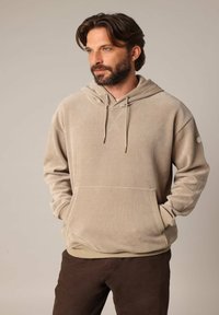 Beige corduroy hoodie met een voorzak, trekkoordkap en ribgebreide manchetten. Model draagt een broek in bruin met een gladde textuur.