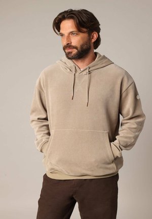 Sweat-shirt en velours côtelé beige avec une poche avant, une capuche à cordon de serrage et des poignets côtelés. Le modèle porte un pantalon marron au tissu lisse.