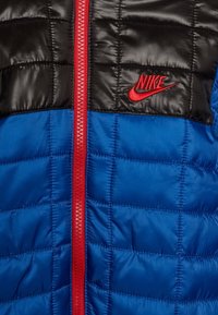 Puffatakki, jossa on musta tikkikangas ylhäällä ja kirkkaan sininen alaosa, punainen vetoketju ja punainen brodeerattu Nike-logo rinnassa.