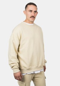 Felpa beige realizzata in un tessuto morbido, con colletto a girocollo, spalle cadenti e polsini a costine. Indossata sopra una camicia bianca e pantaloni abbinati.