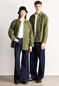 Χακί πράσινα μπουφάν με κλείσιμο με κουμπιά και τσέπες μπροστά, φορεμένα πάνω από λευκά πουκάμισα. Σκούρα, φαρδιά τζιν παντελόνια ολοκληρώνουν το casual look.