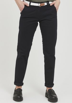 Chino en coton marine avec une coupe droite, présentant des ourlets roulés et des poches latérales. Associé à des mocassins noirs et une ceinture tressée blanche.