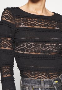 Schwarzes Spitzenoberteil mit horizontalen, strukturierten Streifen, transparentem Stoff, gezackten Kanten und floralen Mustern. Kombiniert mit hochgeschnittenen Jeans.