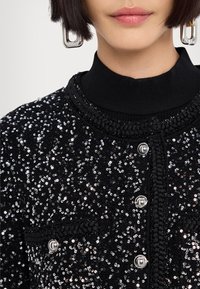 Giacca nera in paillettes con scollatura rotonda, rifiniture intrecciate e chiusura a due bottoni. Presenta un tessuto testurizzato e accenti metallici.