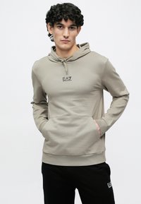 Νεαρός άνδρας με σγουρά μαλλιά, που φοράει μπεζ φούτερ με κουκούλα με το λογότυπο EA7 Emporio Armani και μαύρο παντελόνι με το ίδιο λογότυπο, με τα χέρια στις τσέπες του φούτερ.