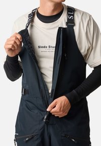 Marineblauwe waterdichte overall met verstelbare banden, rits aan de voorkant en zakken; gedragen over een lichtgekleurde t-shirt. Gestructureerde stof.