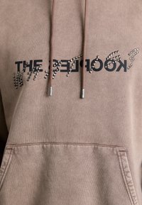 Sweat à capuche marron clair avec texte inversé noir partiellement couvert par des clous pyramidaux argentés sur la poitrine et cordons pendants à l'avant.