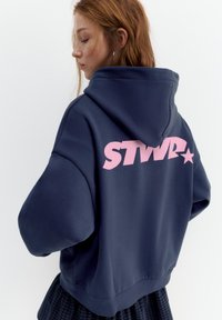 Giovane donna con capelli rossi che indossa una felpa oversize blu marino con scritta rosa "STWR" e stella sul retro, rivolta verso sinistra con gli occhi chiusi.