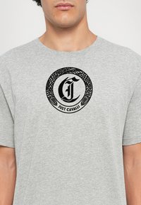 Tricou gri din bumbac cu un logo circular negru care are un motiv cu șarpe și textul „JUST CAVALLI” în centru. Mâneci scurte, decolteu rotund.