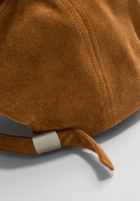 Casquette en daim marron avec une texture lisse, dotée d'un fermoir en métal et d'une lanière réglable pour un meilleur ajustement. Forme arrondie au sommet.