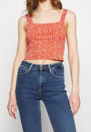 Kobieta w czerwonym, krótkim topie na ramiączkach z białym kwiatowym wzorem i wysokim stanem niebieskich dżinsów, stojąca na jednolitym tle.