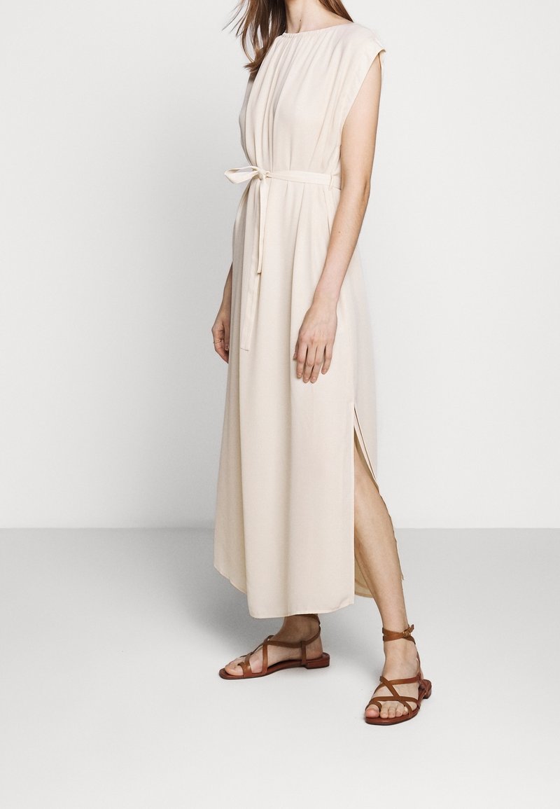 Femme portant une robe midi beige sans manches avec une fente latérale et une ceinture à nouer, associée à des sandales plates marron à brides, debout sur un sol blanc.