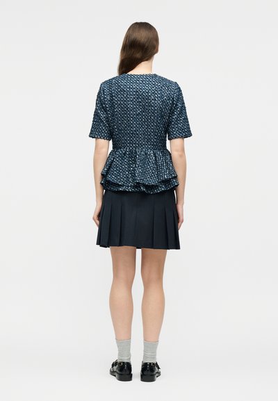 Femme debout de dos portant un haut péplum bleu texturé à manches courtes, une jupe plissée bleu marine, des chaussettes grises et des chaussures noires.