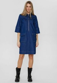 Denimklänning, knälång med korta ärmar, har en dragkedja framtill, två stora fickor och kontrastsöm. Bärs med svarta ankelstövlar.