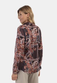 Blusa a maniche lunghe con intricati motivi paisley e geometrici in rosso, blu e bianco su sfondo nero; tessuto leggero e design con collare.