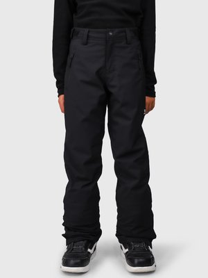 Osoba oblečená v čiernych nohaviciach so zipsom a čiernych teniskách s bielou podrážkou, stojaca pred jednoduchým pozadím.