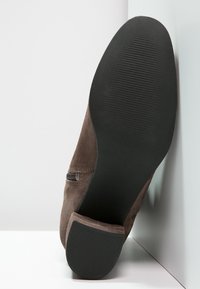 Chaussure en daim marron à bout rond, avec un détail de fermeture éclair sur le côté et une semelle en caoutchouc noir texturé, posée en angle sur un fond clair.