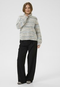 Kobieta w wielokolorowym swetrze z golfem, luźnych czarnych spodniach i butach w czarno-białe paski stoi i uśmiecha się na jednolitym tle.