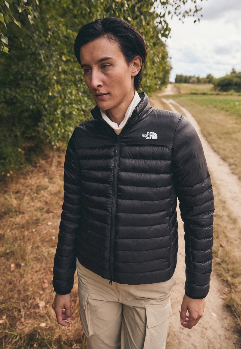 Giacca a vento nera con cuciture orizzontali, chiusura con zip e colletto alto. Indossata sopra una camicia chiara, ambientata in uno sfondo naturale.